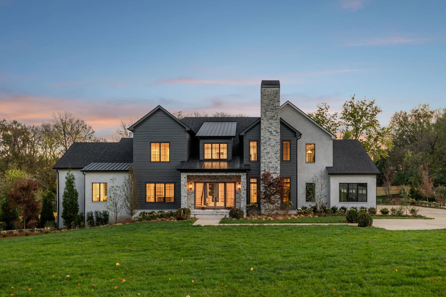 Modern two-story house with dark siding, light stucco, stone chimney, and lit windows at dusk.