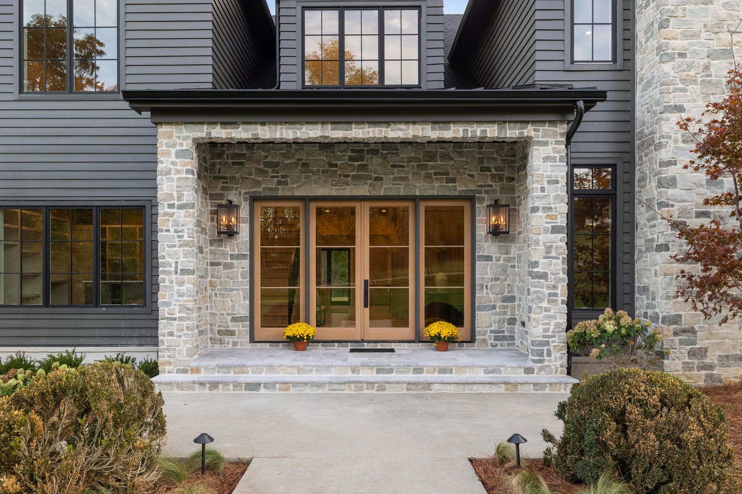 Stone facade house entrance with wooden double doors, windows, lanterns, and yellow flowers.