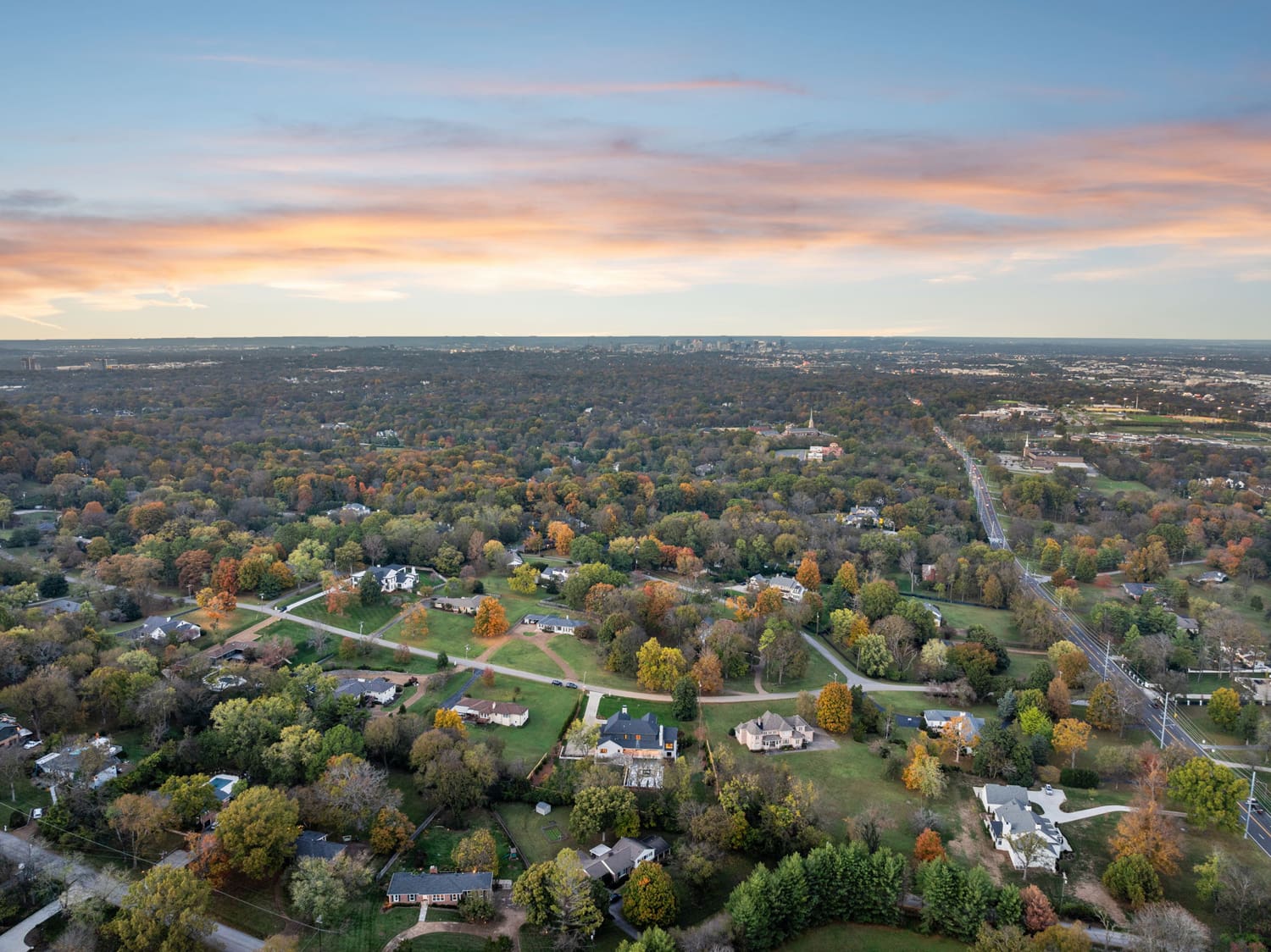 Aerial view of suburban houses amid autumn trees with a road and city skyline at sunset.