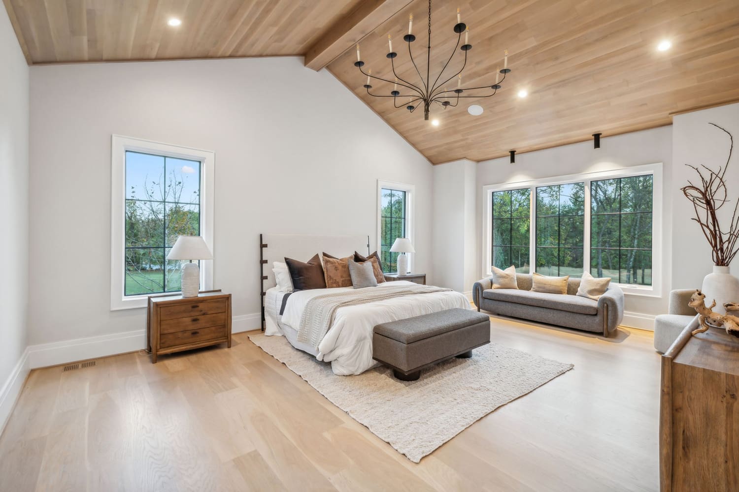Bedroom with bed, gray bench, wooden nightstands, gray sofa, large windows, and wood ceiling.