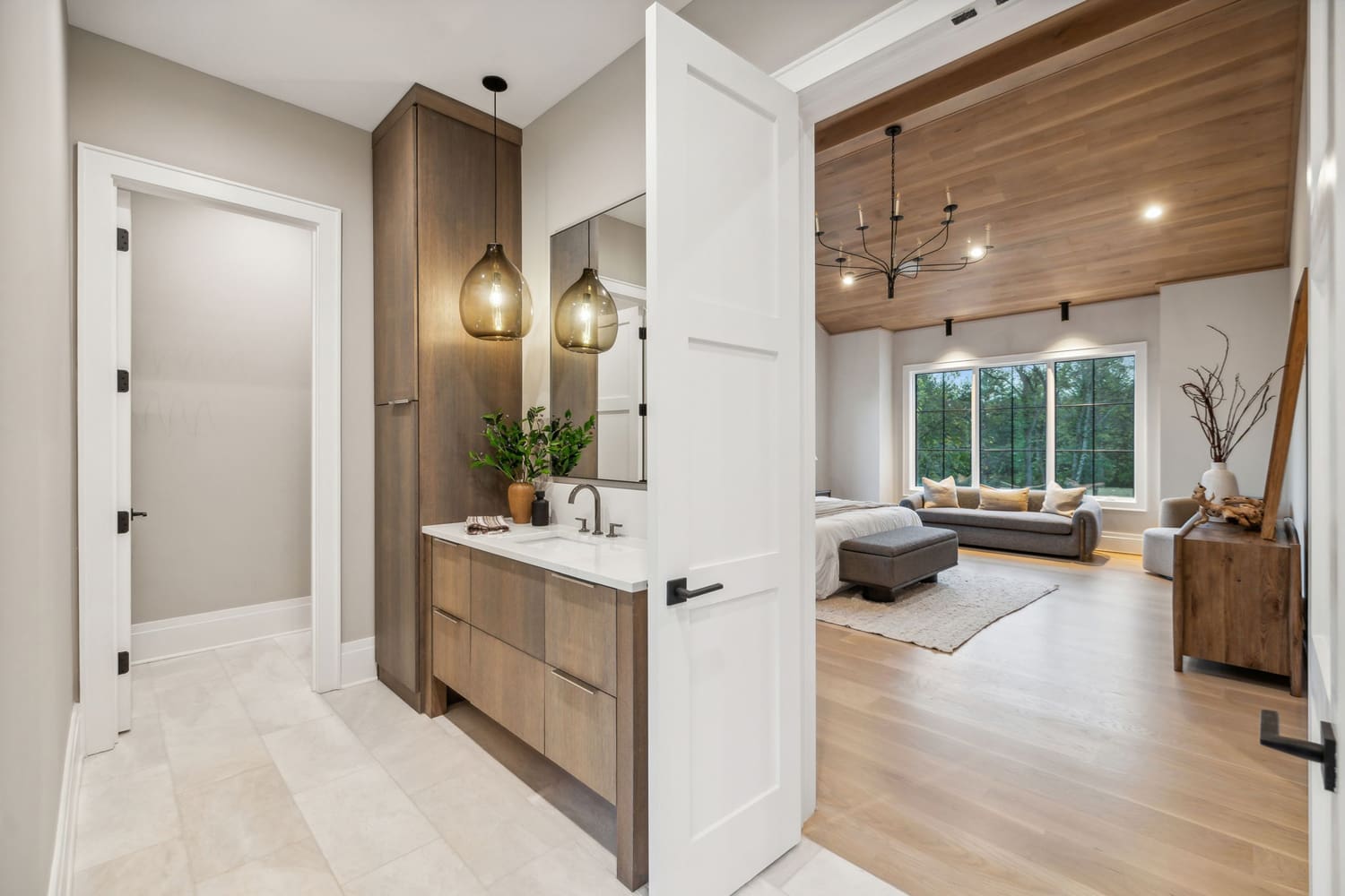Bathroom with wooden vanity and pendant light opens to bedroom with wooden ceiling and large window.