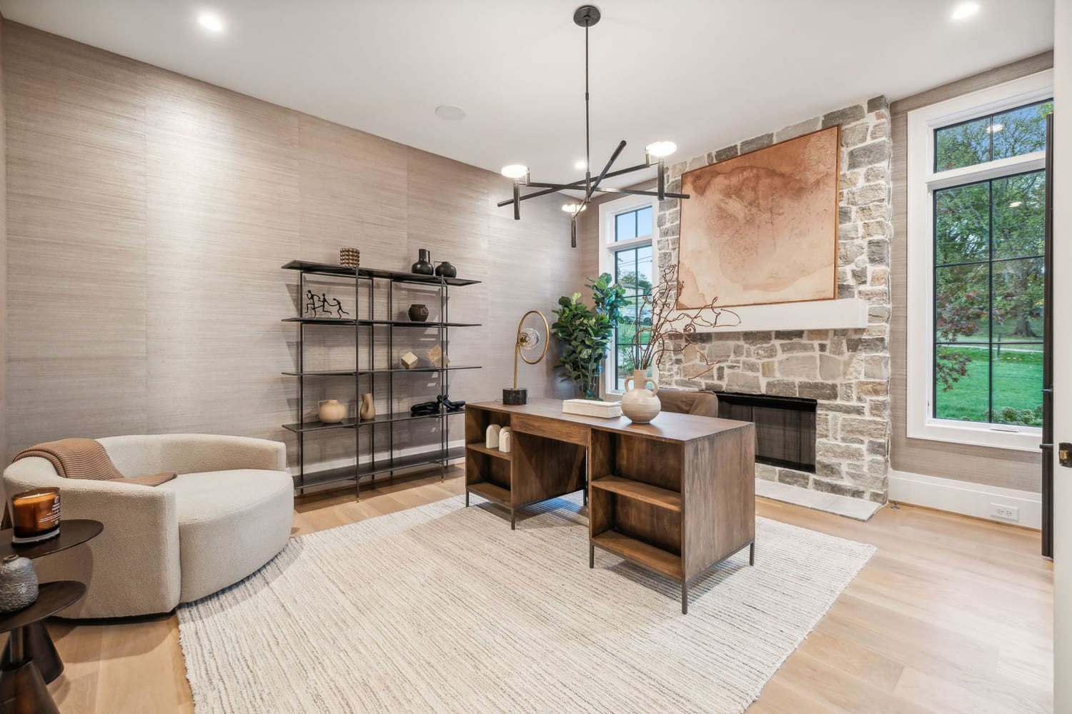 Modern home office with wooden desk, beige chair, metal shelving, stone fireplace, and large windows.