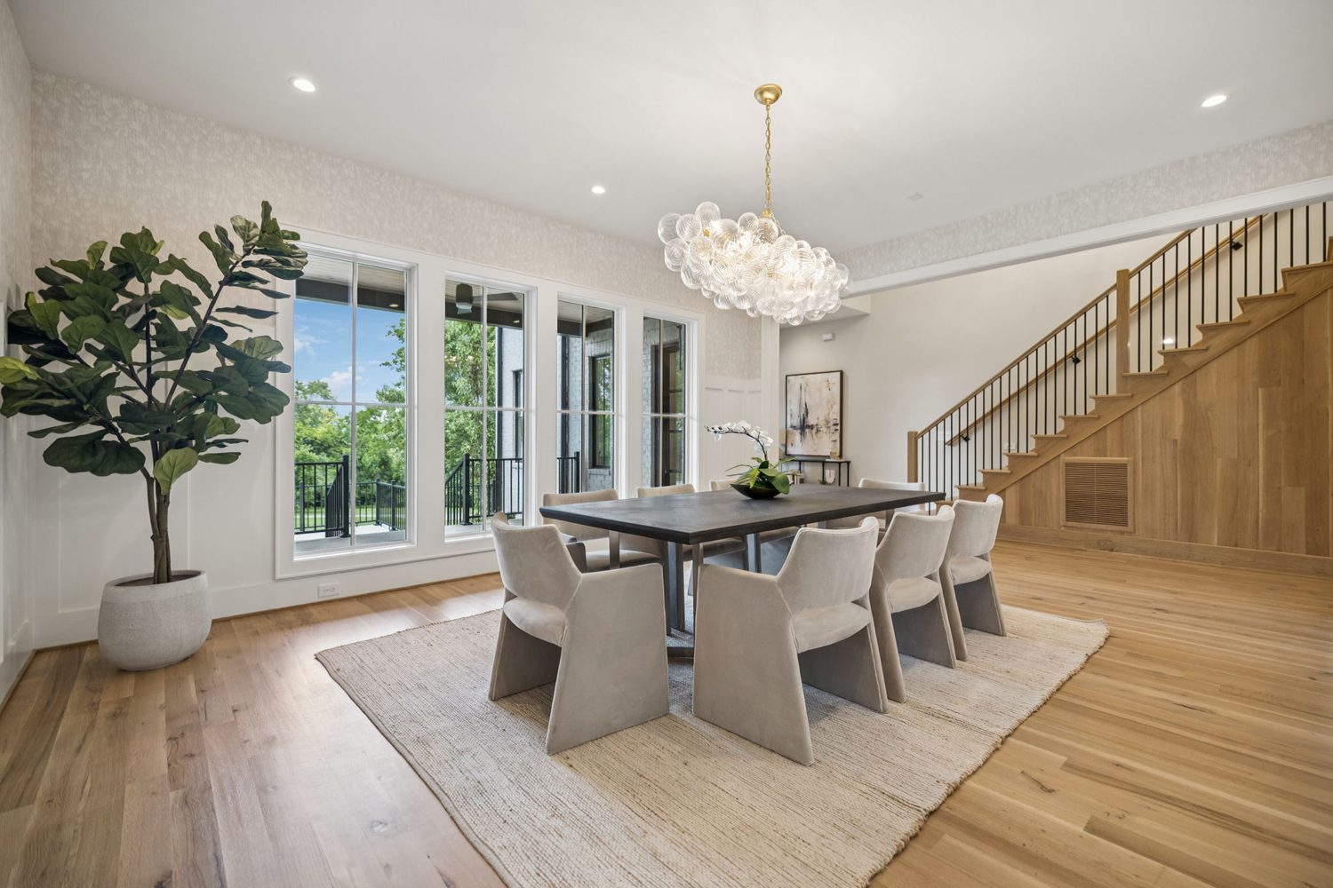 Dining room with rectangular table, eight chairs, large windows, chandelier, staircase, and plant.