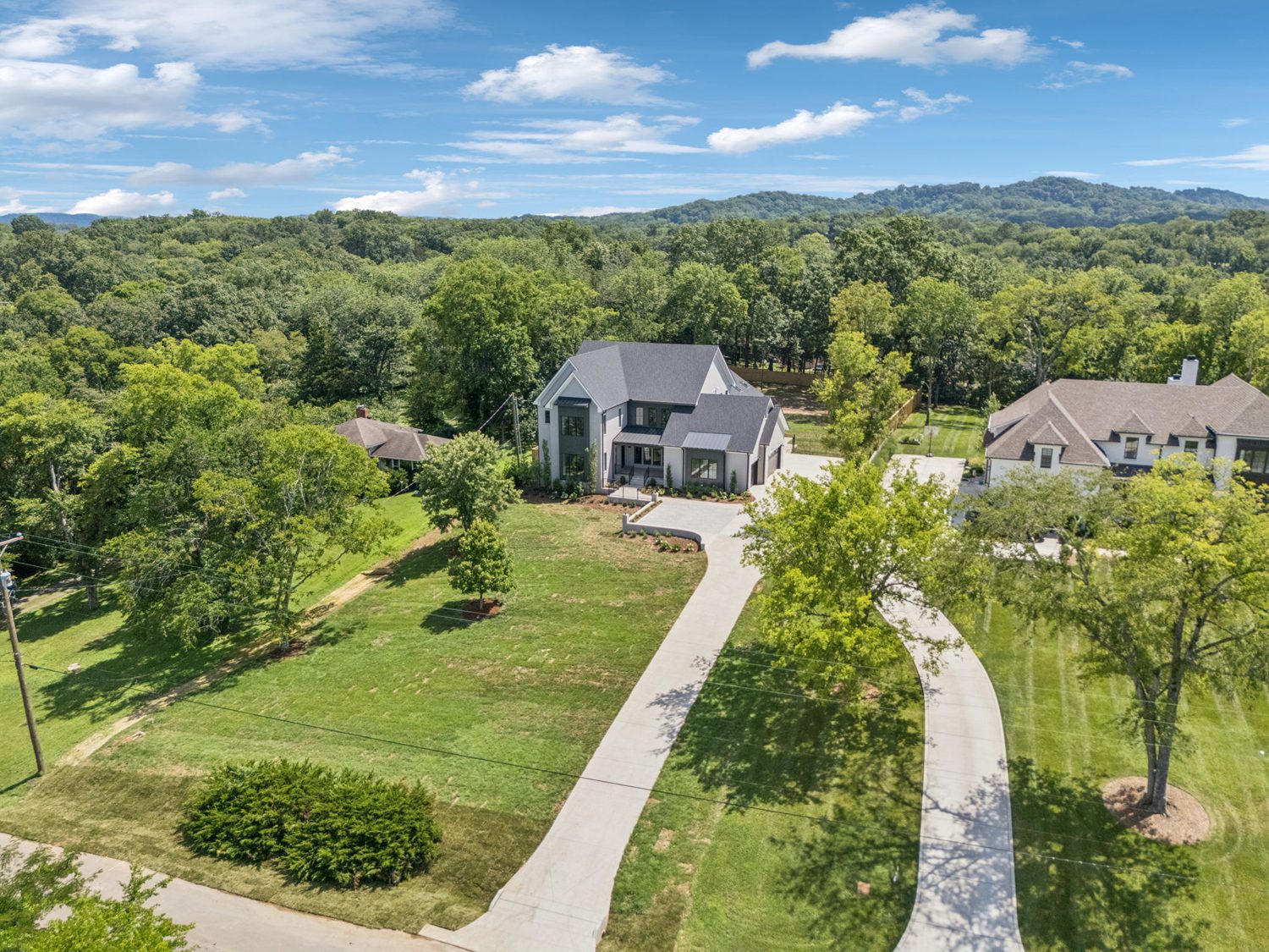 Aerial view of two modern houses with large driveways, lawns, trees, and wooded hills in the background.