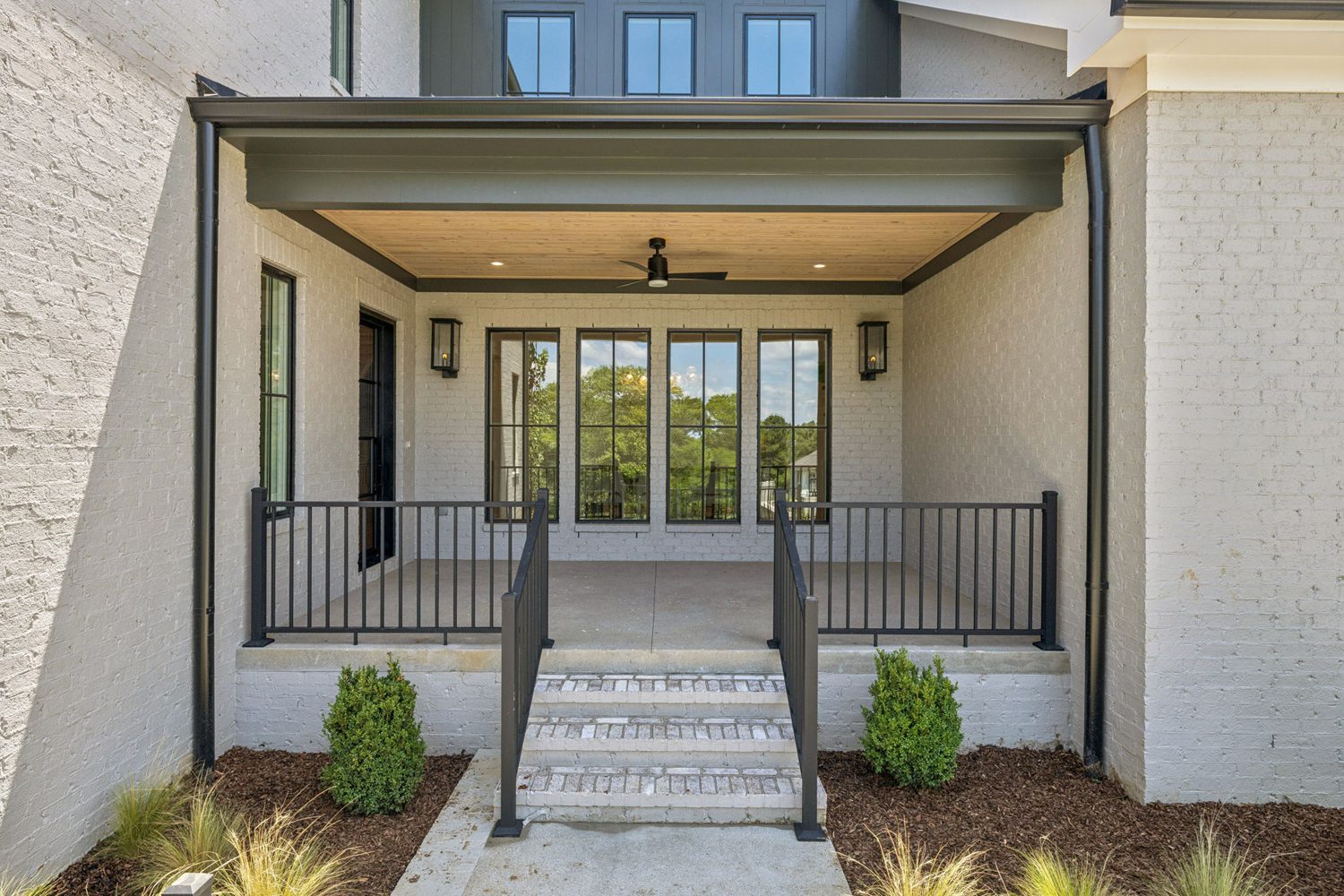 Covered outdoor patio with brick walls, wooden ceiling, ceiling fan, black railings, and large windows.