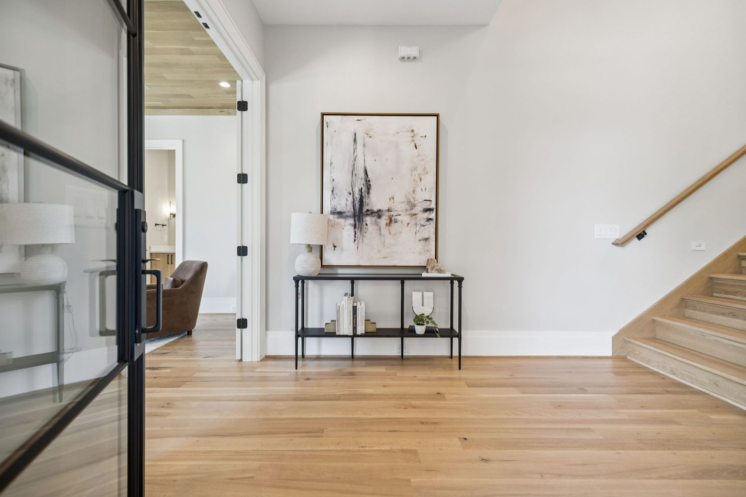 Minimalist interior with light wood floor and stairs, black console table, lamp, abstract painting.