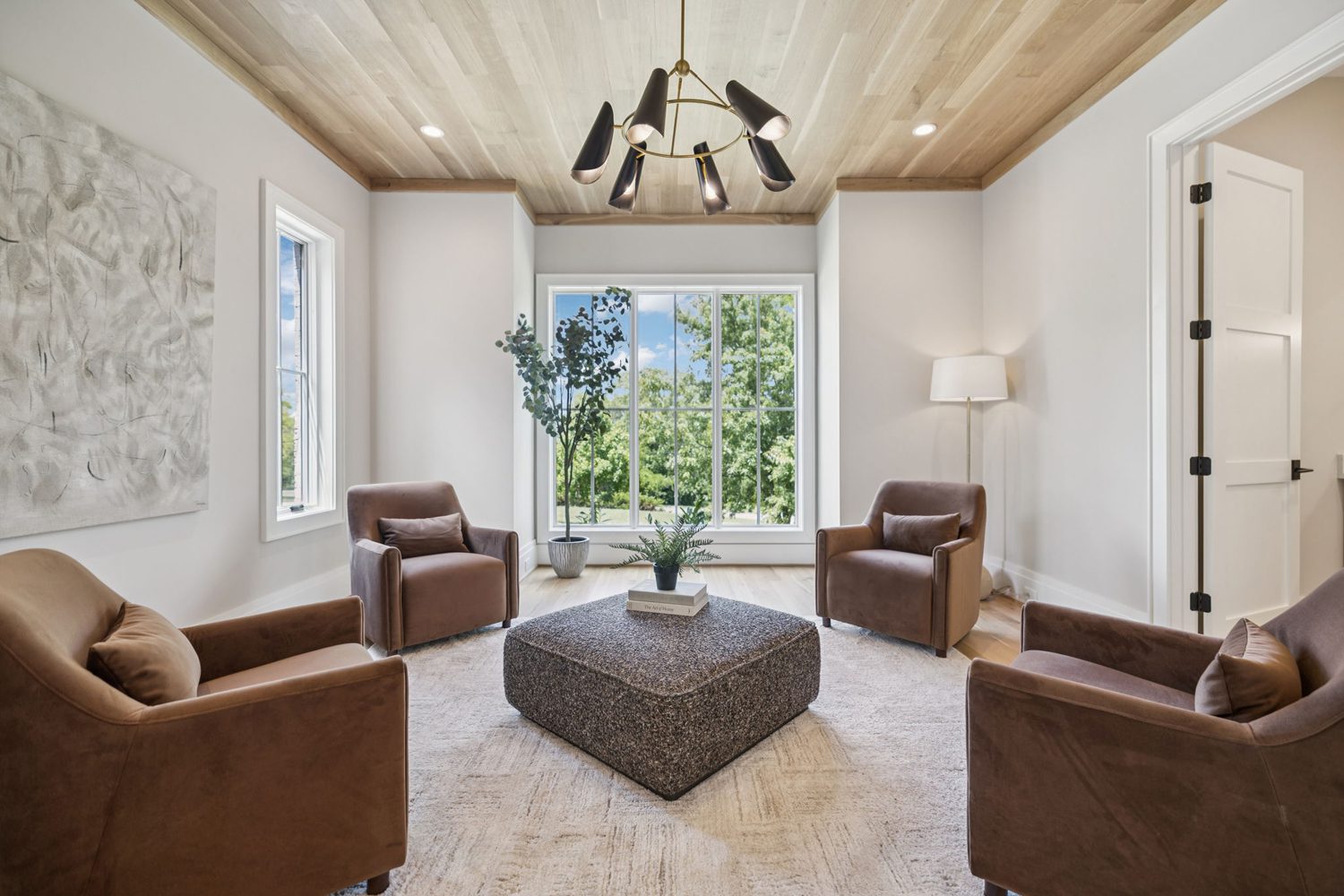 Living room with four brown armchairs around a square ottoman, large window, and wooden ceiling.
