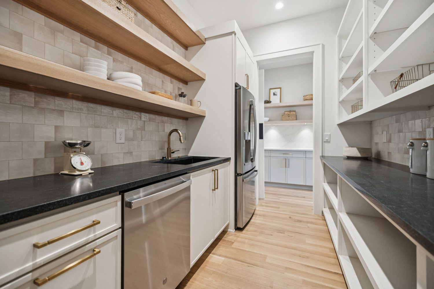 Narrow modern kitchen with light wood flooring, white cabinets, black countertops, and stainless appliances.