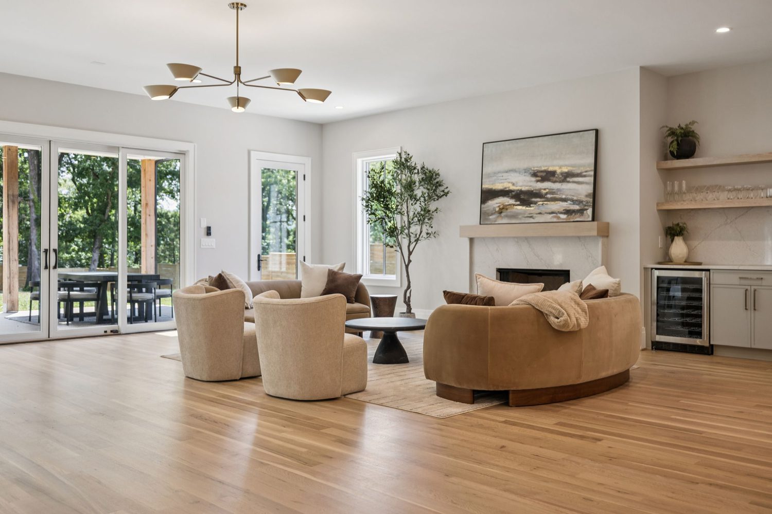 Living room with light wood floors, neutral seats, fireplace, plant, cabinet with wine cooler, and glass doors.