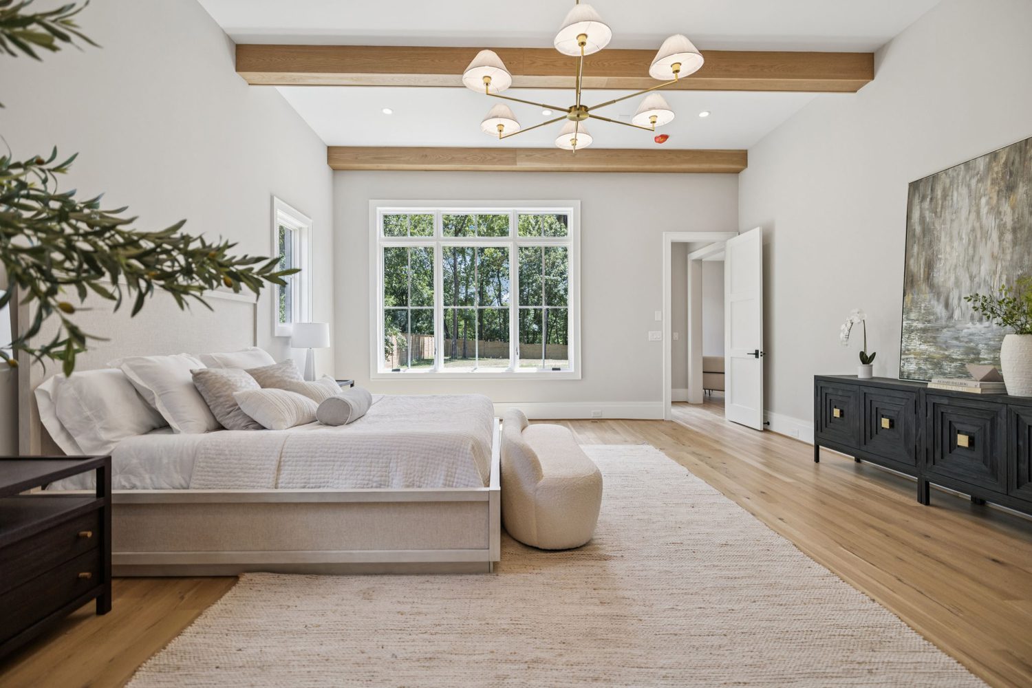Spacious bedroom with large window, wooden beams, light wood floor, bed, bench, dark furniture, and artwork.