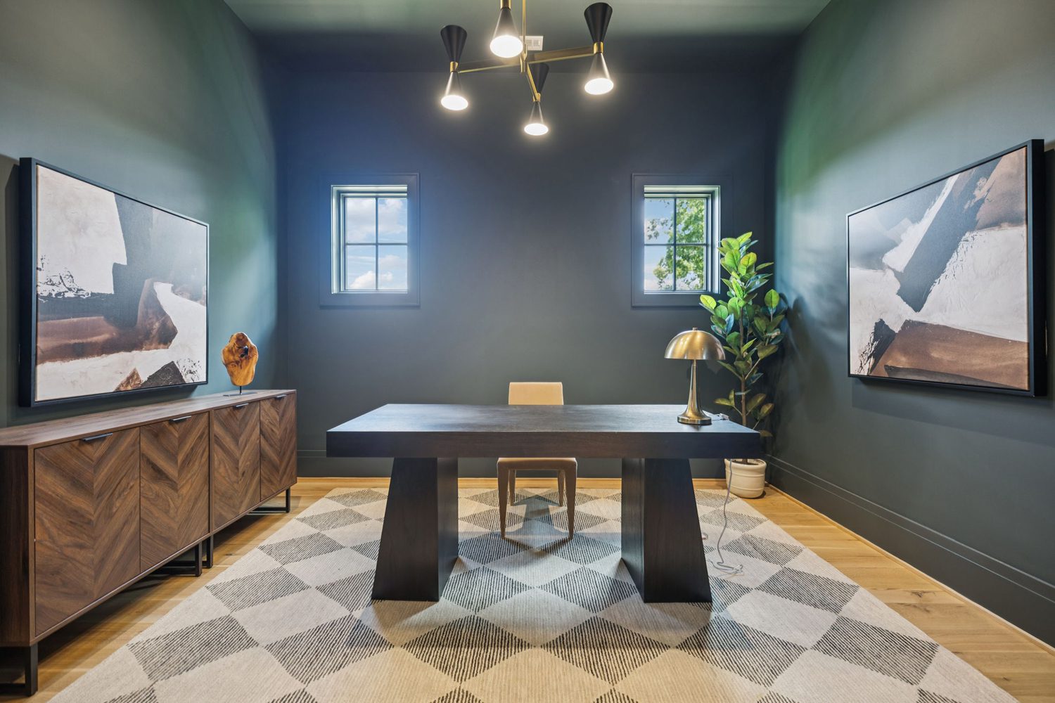 Office with dark gray walls, two windows, large dark desk, tan chair, sideboard, artworks, plant, and chandelier.