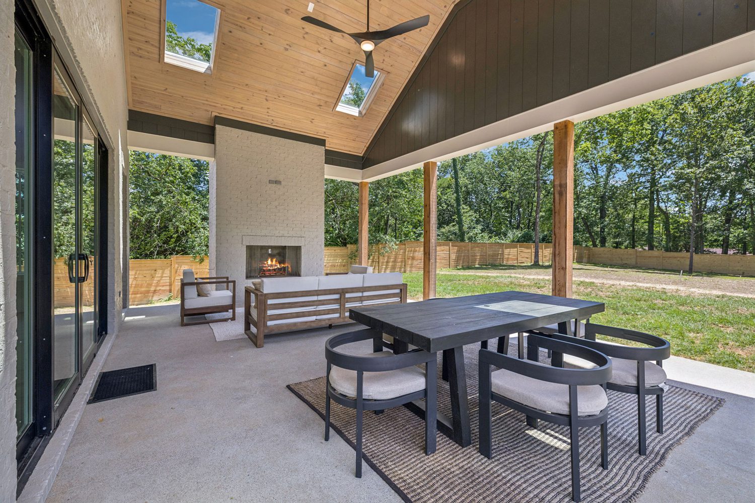 Covered patio with dining table, seating area, fireplace, ceiling fan, skylights, and fenced yard.