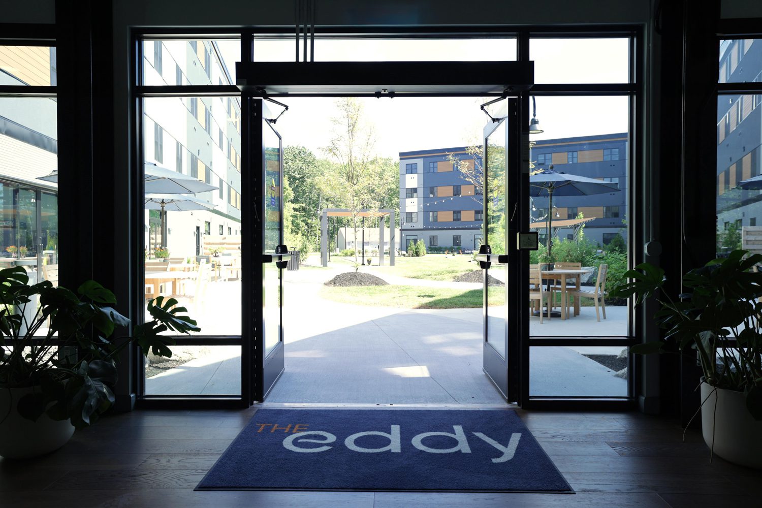 Entrance with open glass doors, welcome mat reading The eddy, plants, courtyard, and modern buildings.