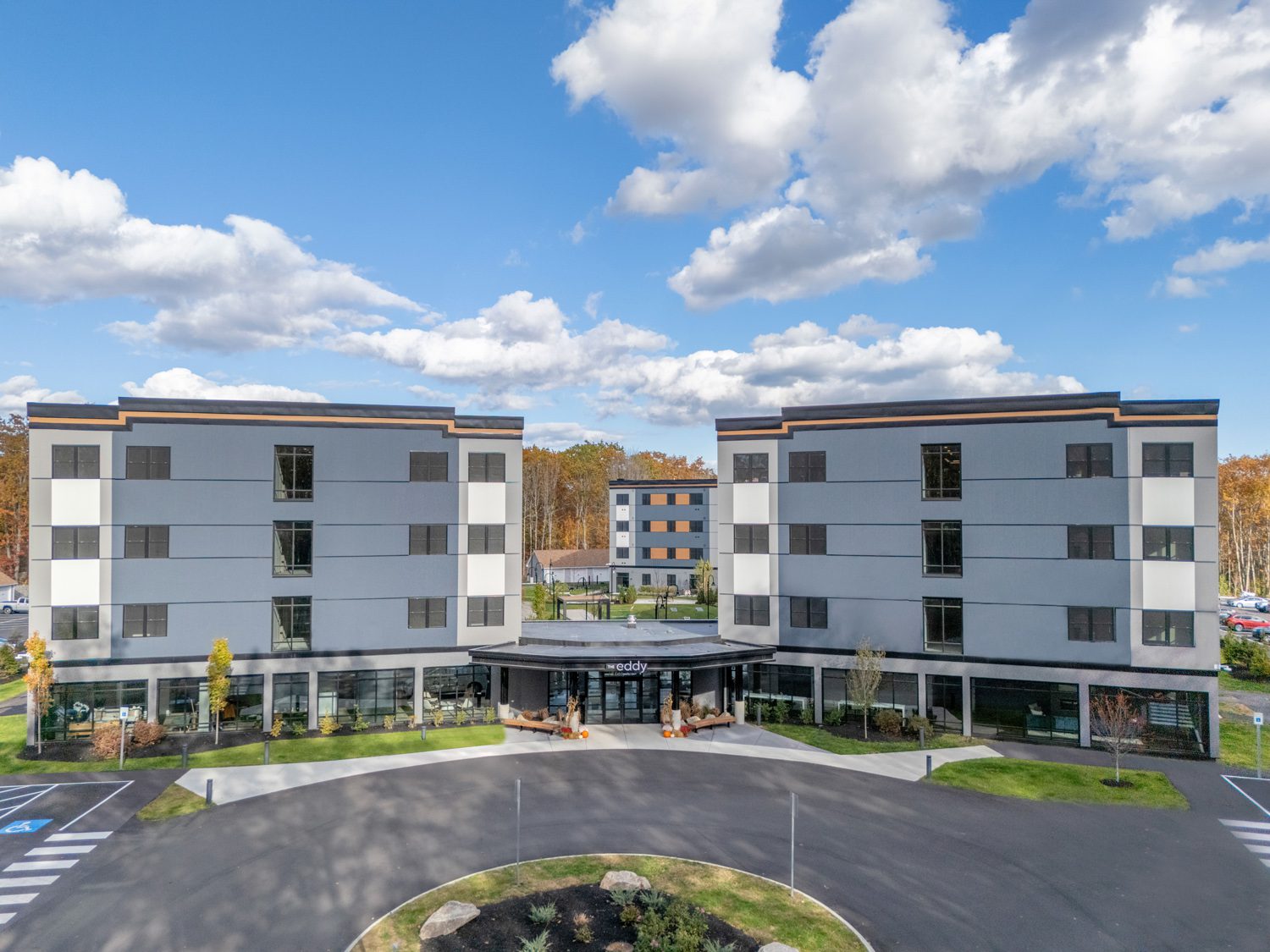 Four-story modern gray building complex with central entrance and landscaped surroundings under blue sky.
