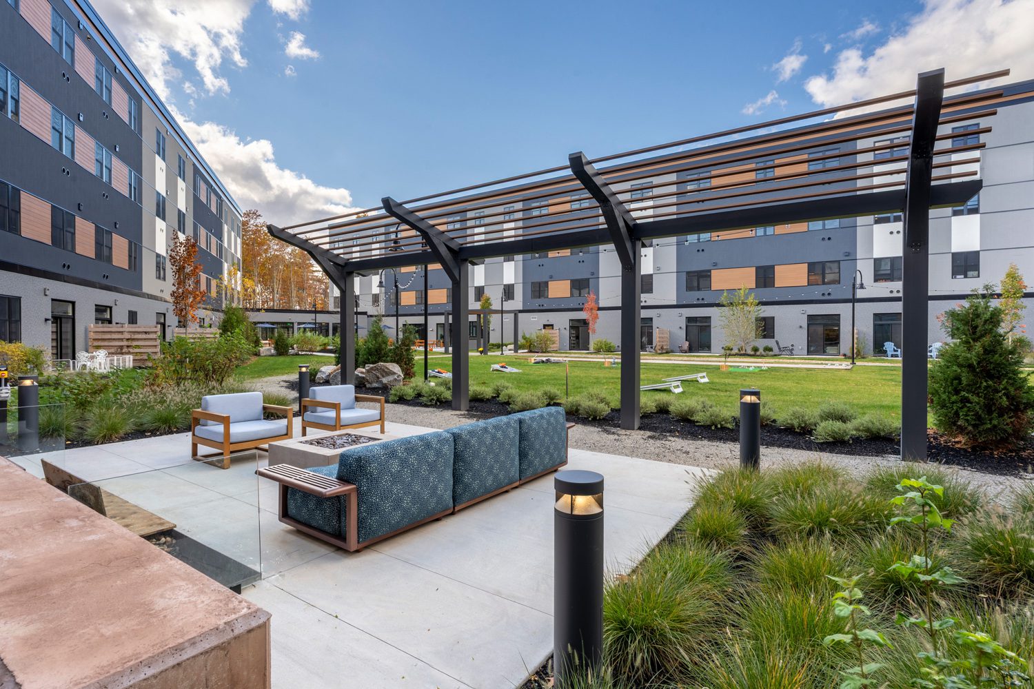 Outdoor courtyard with modern seating, fire pit, pergola, grassy area, and cornhole boards.