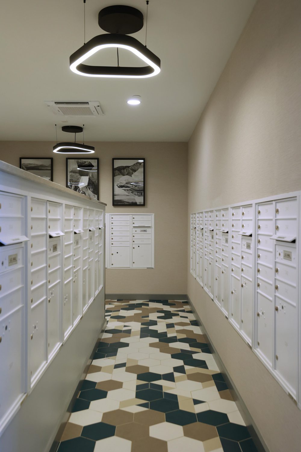 Hallway with white mailboxes on both sides, geometric tile floor, modern ceiling lights, and black-and-white framed photos at the end.