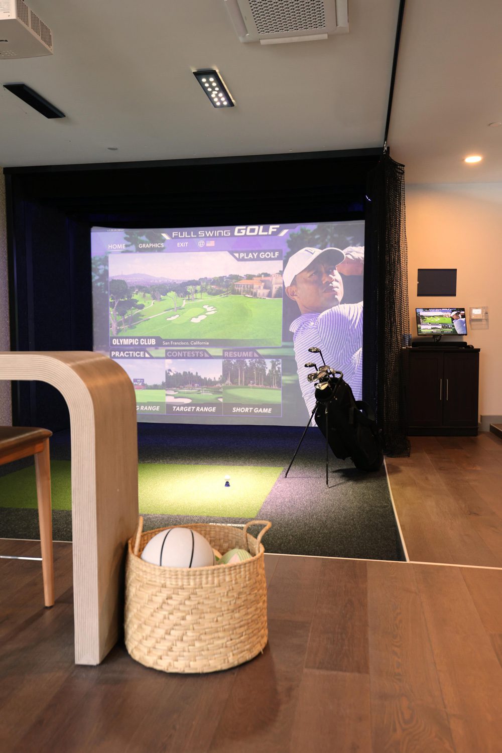 Indoor golf simulator showing a golf game screen, golf clubs on stand, and basket with balls