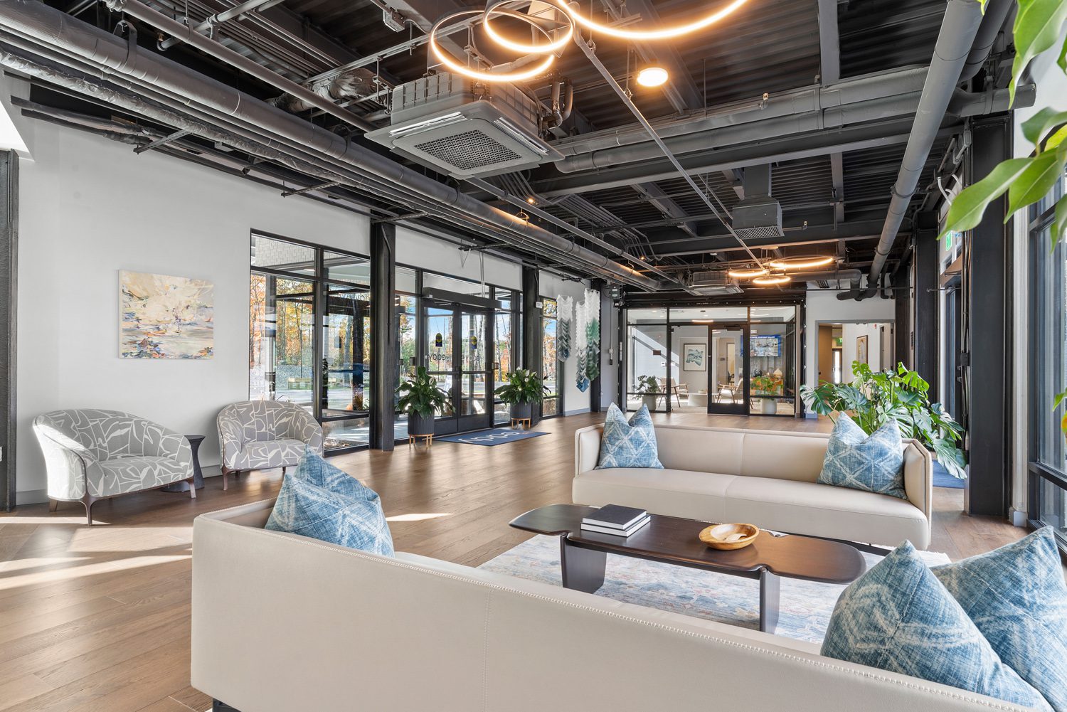 Modern lobby with exposed ceiling, light sofas, blue pillows, armchairs, plants, and glass doors.