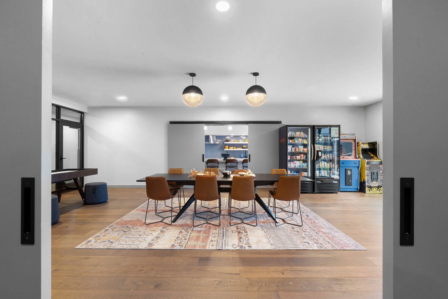 Communal room with large table, brown chairs on patterned rug, vending machines, arcade games, foosball table, and kitchen area.