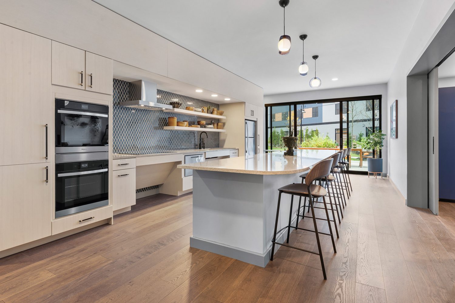 Modern kitchen with light wood cabinets, large island, bar stools, ovens, and sliding glass doors.