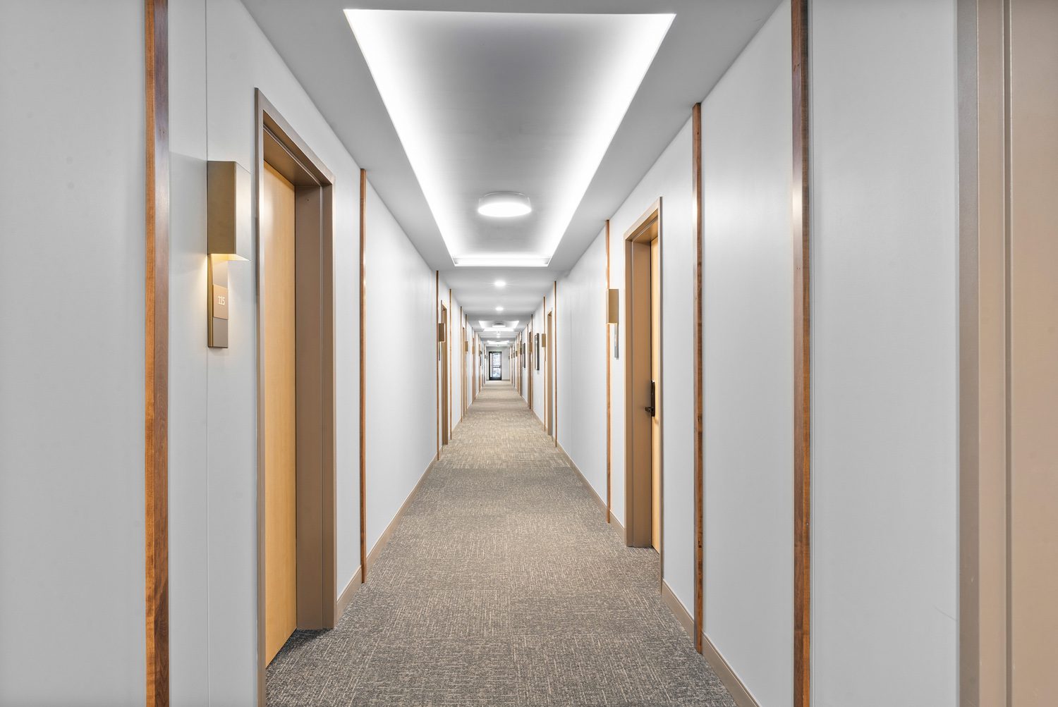 Long, well-lit hallway with closed wooden doors, white walls, and gray carpet flooring.