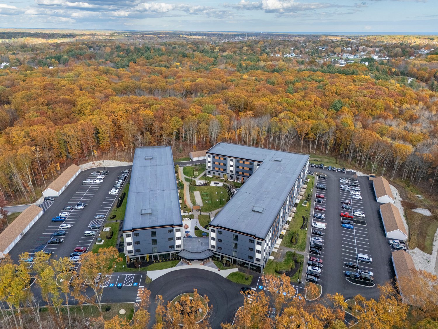 Aerial view of a U-shaped modern building with parking lots near a forest with autumn leaves.